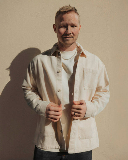 A man wearing The Workwear Overshirt in cream over a white t-shirt, standing outdoors in front of a beige wall. The image highlights the relaxed fit and patch pockets of the overshirt.